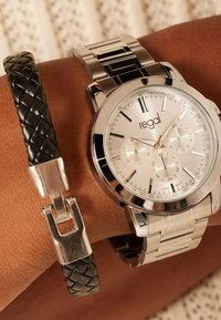 Silver stainless steel watch with a round face and tachymeter. Features a white dial, black accents, and a black braided leather bracelet.