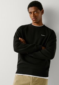 Sweatshirt preto com punhos e bainha canelados, gola redonda e pequeno logótipo branco no peito. Usado sobre uma t-shirt branca. Textura suave.