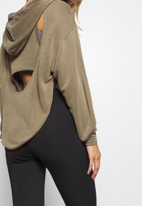 Olivgrön oversized hoodie med utskärning på baksidan, långa ärmar och textilstruktur, tillsammans med svarta leggings.