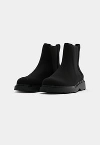 Botas de tornozelo pretas feitas de material liso, com bico arredondado e painéis laterais elásticos. Sola grossa preta com ranhuras para tração.