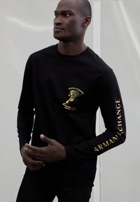 Melnā ar garām piedurknēm krekls ar zelta grafisko dizainu uz krūtīm un piedurknēm. Gluda tekstūra, vaļīgs siluets, ar logo un motivējošu tekstu.