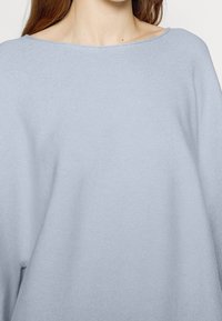 Pull bleu clair, ample, avec un col large et un tissu en maille texturée. Les manches sont larges, créant une silhouette décontractée.