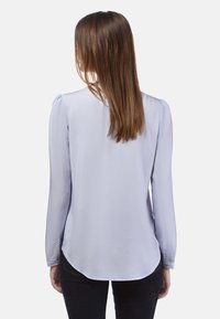 Lichtblauw blouse met lange mouwen, een gladde textuur, een ronde onderkant en puffy schouderdetails. Draagt met donkere jeans.
