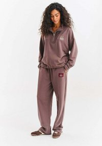 Brązowa bluza z zamkiem na ćwiartkę i logo, zestawiona z pasującymi spodniami z małą naszywką z logo, wykonana z miękkiego, teksturowanego materiału.