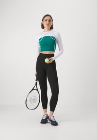 Dugi rukav bijela bluzica preko zelenog kratkog topa, crne rebraste tajice, drži lopticu za tenis i reketa, nosi tamnoplave atletske tenisice.