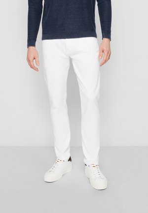 Mann trägt schmale weiße Hose, weiße Sneaker und einen dunkelblauen Pullover mit langen Ärmeln und steht vor einem einfarbigen Hintergrund.