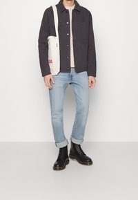 Sort jakke med knapper, cremefarvet t-shirt, lyseblå jeans med manchetter, sorte snørebåndsstøvler og en beige tote-taske med rød tekst.