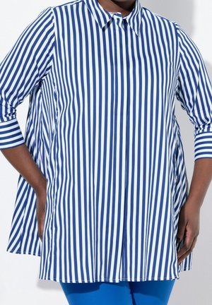 Osoba v modro-bíle pruhované košili s dlouhými rukávy a límečkem a modrých kalhotách, ruce opřené o boky.