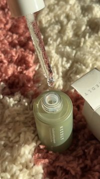 Contagocce che rilascia siero sopra una bottiglia verde aperta con etichetta "NEEDLY" su una superficie rosa e bianca texturizzata con confezione parzialmente visibile.