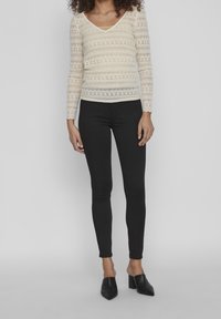 Lace långärmad topp i krämfärg med V-ringning och vågformade kanter, kombinerad med svarta slim-fit byxor och svarta spetsiga loafers.