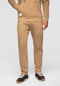 Homme portant un pantalon de survêtement beige et un sweat à capuche assorti, les mains dans les poches, baskets noir et blanc, debout sur un fond neutre.