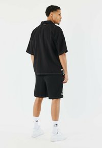 Camisa negra de manga corta acanalada y pantalones cortos a juego, con costuras y etiquetas de logo en contraste. Conjunto estilizado con zapatillas blancas y calcetines.