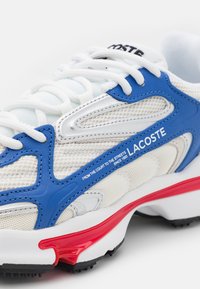 A Lacoste sportcipő fehér hálós felsőrésszel rendelkezik, kék szintetikus kiegészítőkkel, ezüst részletezéssel és piros gumitalppal. Az oldalon szöveg található.