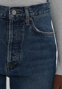 Gros plan d'un jean en denim bleu avec bouton fermé, poche avant visible, passants de ceinture et partie de la manche d'un pull gris côtelé ainsi qu'une main.