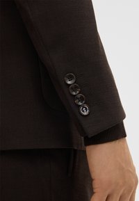 Donkerbruine blazer mouw met vier zwarte knopen, voorzien van een textuurstof en geribbelde manchetten. De tailoring bevat subtiele details en stiksels.