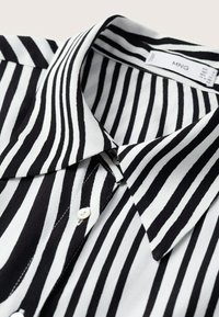 Chemise à rayures verticales noir et blanc avec un col pointu et une étiquette de marque visible à l'intérieur du cou.