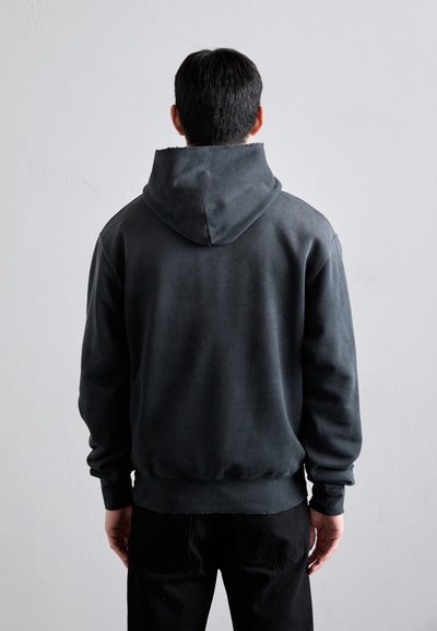 Sudadera gris oscuro con una gran capucha, puños y dobladillo acanalados, que presenta una textura suave y un ajuste relajado. Pantalones negros parcialmente visibles.