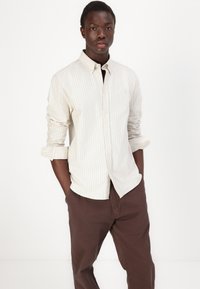Chemise rayée à boutons crème et beige clair, en tissu mélangé de coton, manches longues, assortie à un pantalon marron foncé.