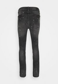 Jean skinny noir avec poches arrière et motif éclaboussures de peinture blanche, vu de dos sur un fond uni.