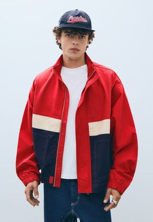 Junger Mann trägt rot-weiß-navy Jacke, weißes Hemd, navy Cap mit "fearless" in rot und dunkle Jeans, steht vor schlichter Hintergrund.