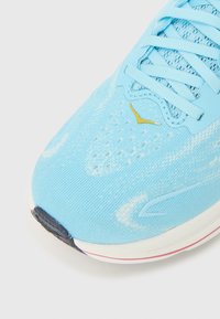 Ανοιχτό μπλε αθλητικό παπούτσι με υφασμάτινο επάνω μέρος με υφή πλεκτού, επίπεδα κορδόνια και κίτρινη λεπτομέρεια κοντά στα κορδόνια. Λαστιχένια σόλα με κυρτό σχεδιασμό.