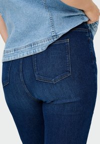 Artimas planas, kuriame matyti asmuo, vilkintis tamsiai mėlynus džinsus ir šviesiai mėlyną džinsinį marškinį, ranka įsikišta į džinsų kišenę.