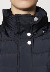 Marineblauwe pufferjack met een hoge kraag, zilveren drukknoopsluiting, gequiltde textuur en ritssluiting voor extra warmte.