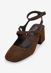 Zapato de tacón cuadrado de ante marrón con correa en el tobillo, detalle entrecruzado y punta cuadrada. Presenta un forro interior suave y una suela texturizada.