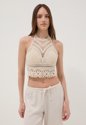 Joven mujer que lleva un top corto de ganchillo color crema con cuello halter combinado con pantalones sueltos color crema con cordón, sobre un fondo liso.