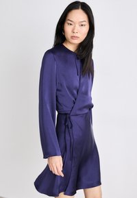 Ljubičasta satenasta haljina omotnica s dugim rukavima, detaljem vezanja u struku i lepršavom suknjom. Glatka tekstura i elegantno pada.