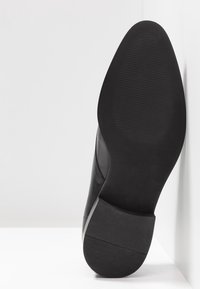 Chaussure en cuir noir avec une finition lisse, dotée d'un bout arrondi et d'une semelle texturée noire. La hauteur du talon est modérée avec des détails de couture subtils.