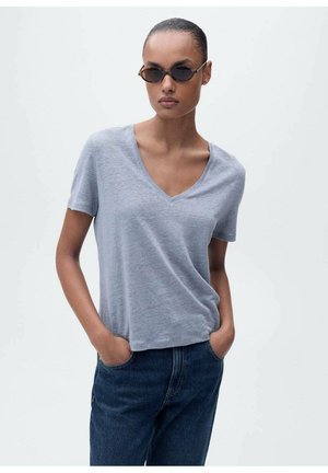 Szara, krótko rękawowa koszulka V-neck wykonana z miękkiego materiału, w połączeniu z ciemnoniebieskimi dżinsami. Model nosi wąskie okulary słoneczne, ręce w kieszeniach.