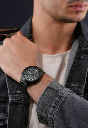 Black wristwatch with a textured face, silver accents, and a metal strap. Worn on a wrist, beside a gray denim jacket and white shirt.