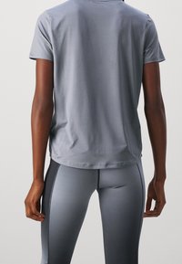 Persona vistiendo una camiseta corta gris holgada y leggings en degradado gris, de pie con los brazos relajados a los lados.