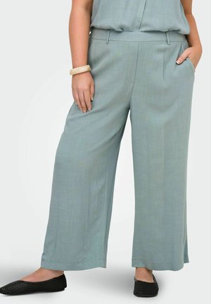 Kobieta ubrana w jasnoniebieskie spodnie o szerokich nogawkach, dopasowany bezrękawnik, beżową bransoletkę i czarne, plecione baleriny, stoi z jedną ręką w kieszeni.