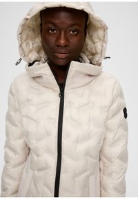 Helle beige Pufferjacke mit diagonaler Steppung, schwarzem Reißverschluss und verstellbarem Kapuzen. Verfügt über Seitentaschen und ein Logo-Detail am Ärmel.