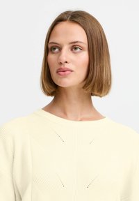 Pletený svetr v měkké krémově žluté barvě, s ribovanou texturou a šikmými perforacemi. Kulatý výstřih a volný střih.
