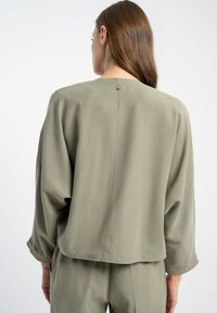 Oliwkowa bluzka o luźnym kroju, z okrągłym dekoltem, długimi rękawami, małym guzikiem z tyłu i gładką fakturą.