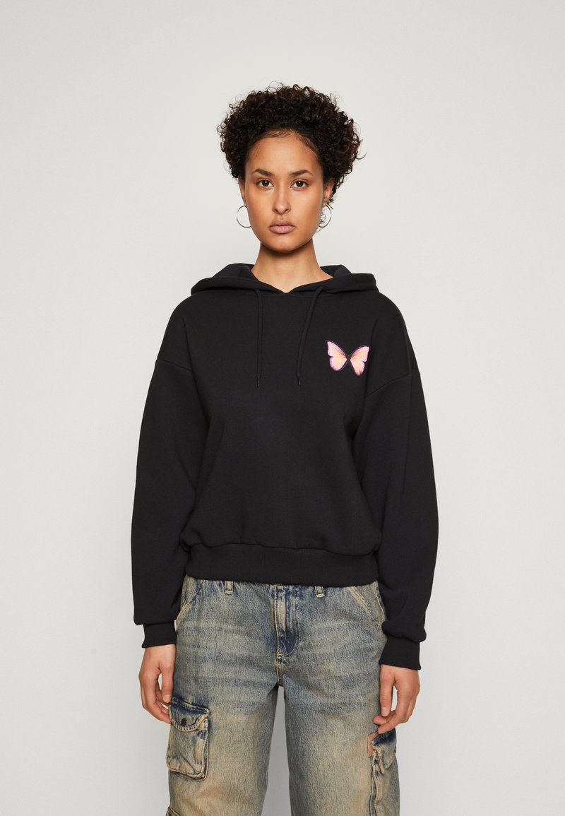 Žena s kratkom kovrčavom kosom nosi crnu hoodicu s ružičastim leptirím amblem i izblijedjele cargo traperice na jednostavnoj svijetloj pozadini.