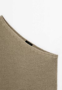 Gros plan d’un tissu beige tricoté avec une encolure diagonale et une petite étiquette noire de vêtement sur fond blanc.