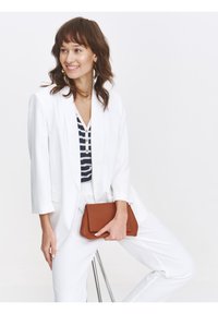Biała marynarka o luźnym kroju, zestawiona z granatowo-paskowanym topem. Modelka trzyma brązową torebkę o teksturze; jasne tło podkreśla detale stroju.