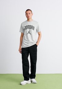 Mand står med den ene hånd i lommen iført en grå Jordan T-shirt, sorte bukser og hvide sneakers på en lys grøn gulv foran en hvid væg.