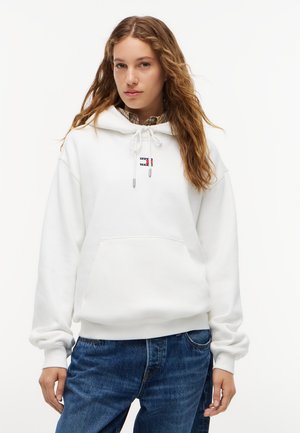 Kvinde iført en hvid hættetrøje med et lille flaglogo på brystet og blå jeans, stående mod en ensfarvet hvid baggrund.