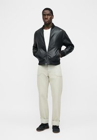 Bărbat purtând o jachetă bomber din piele neagră, tricou alb, pantaloni bej întorși la tiv și adidași negri, stând cu mâinile în buzunare.