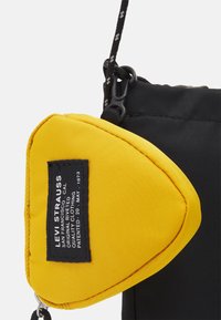 Pochette en tissu noir avec une poche triangulaire jaune, ornée d'une étiquette noire avec texte blanc et d'une sangle assortie.