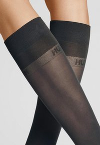 Schwarze Oberschenkelstrümpfe mit einem strukturierten oberen Rand. Die Strümpfe sind transparent mit einem glatten Finish und verfügen über ein geprägtes Logo am Bund.