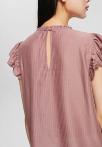 Lyserød bluse med tekstureret stof, flæseærmer i kort længde og en nøglehulsåbning bagpå med knaplukning.