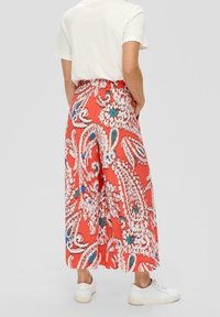 Rote weit geschnittene Hosen mit Paisleymuster aus strukturiertem Stoff, elastischem Bund und weißen Sneakers. Sichtbare Rückansicht.
