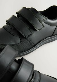 Schwarze Schuhe mit zwei Klettverschlüssen, glattem Leder und strukturiertem Mesh-Material, runder Zehenform und einer Gummisohle mit dezenten Nahtdetails.