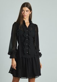 Abito nero con maniche lunghe in tessuto trasparente, colletto arricciato e pattina frontale, con bottoni. Tessuto morbido e design a strati.